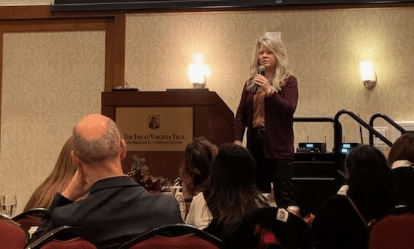 Tracy Smith is holding a microphone and speaking to a group of people in a large conference room.