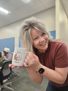 A smiling woman holding a coffee mug with a graphic design, inside a classroom or meeting space.